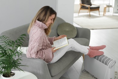 Photo of Happy young woman with book warming feet on electric heater at home