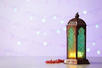 Photo of Muslim lamp Fanus with prayer beads and space for design on blurred lights background