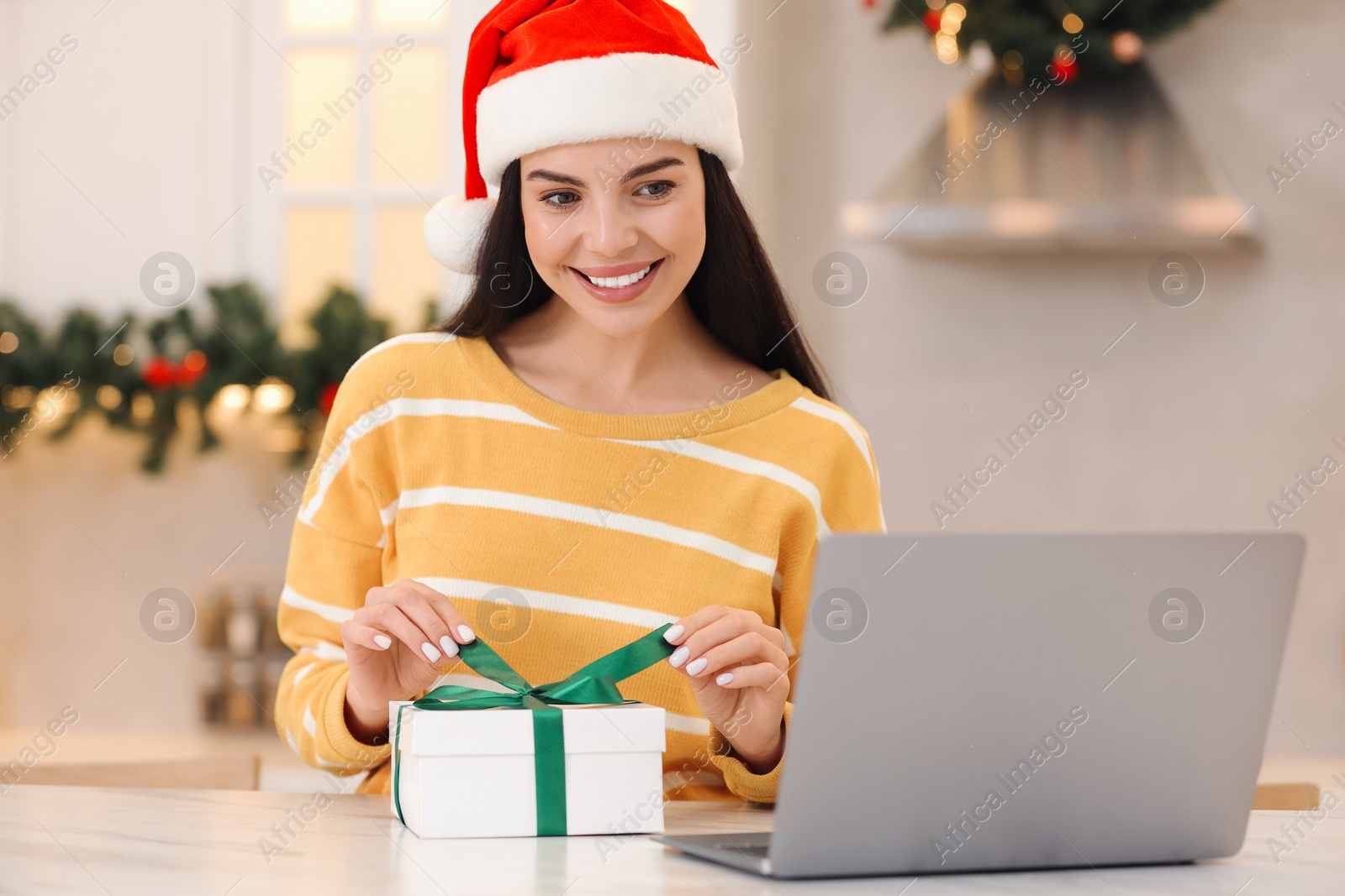 Photo of Celebrating Christmas online with exchanged by mail presents. Smiling woman opening gift box during video call on laptop at home