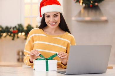 Celebrating Christmas online with exchanged by mail presents. Smiling woman opening gift box during video call on laptop at home