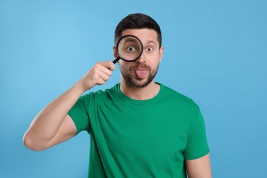 Photo of Man looking through magnifier glass and showing tongue on light blue background
