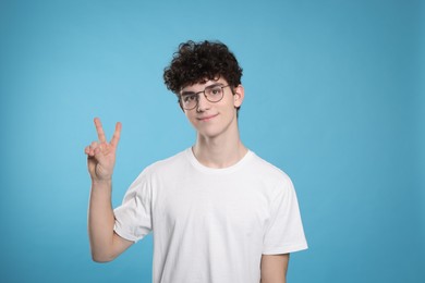 Portrait of cute teenage boy wearing glasses and showing peace gesture on light blue background