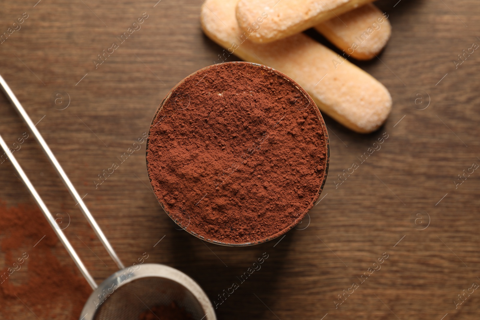 Photo of Delicious tiramisu in glass, biscuits and sieve with cocoa powder on wooden table, flat lay