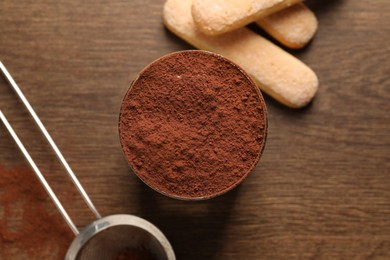 Delicious tiramisu in glass, biscuits and sieve with cocoa powder on wooden table, flat lay