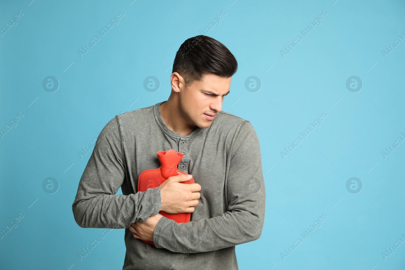 Photo of Man using hot water bottle to relieve chest pain on light blue background