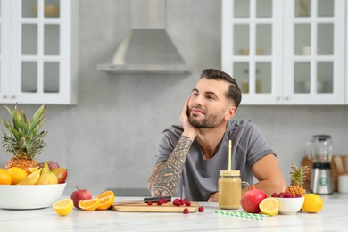 Photo of Handsome man with delicious smoothie and ingredients at white table in kitchen. Space for text