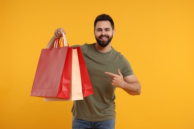 Photo of Happy man pointing at many paper shopping bags on orange background