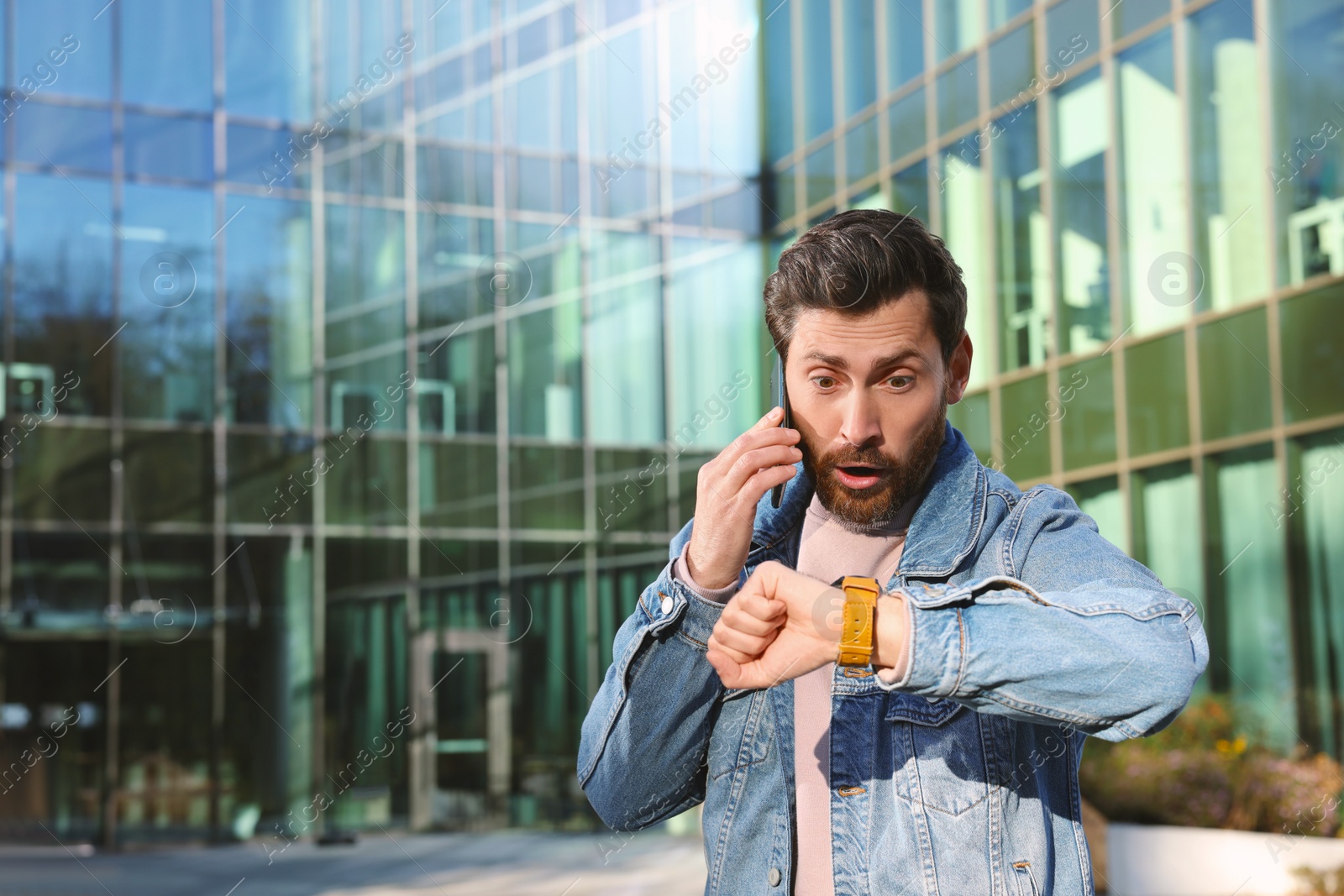 Photo of Emotional man talking on smartphone and looking at watch near building. Being late concept