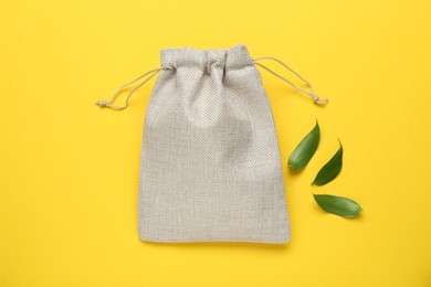 Photo of Burlap bag and green leaves on yellow background, flat lay