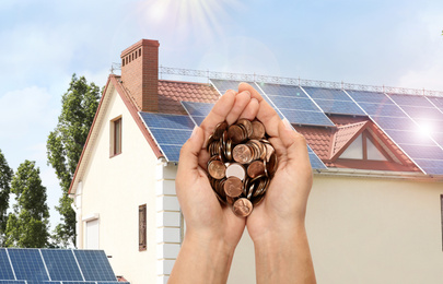 Image of Woman holding coins against house with installed solar panels. Renewable energy and money saving