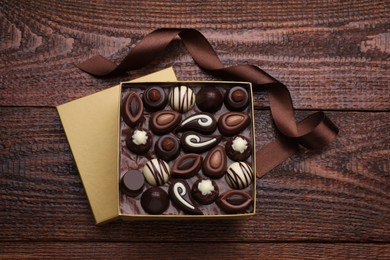 Open box of delicious chocolate candies and brown ribbon on wooden table, flat lay