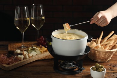 Photo of Woman dipping piece of bread into fondue pot with melted cheese at wooden table, closeup