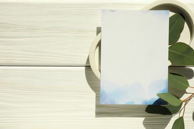 Photo of Blank invitation card and green leaves on white wooden table, top view. Space for text