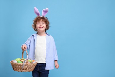 Happy boy in cute bunny ears headband holding wicker basket with Easter eggs on light blue background. Space for text