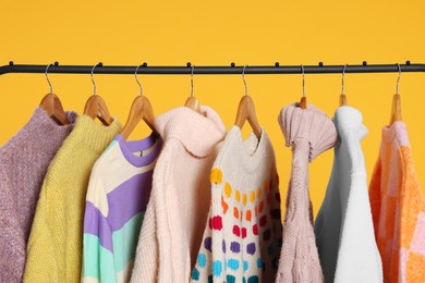 Rack with stylish women's sweaters on wooden hangers against orange background, closeup