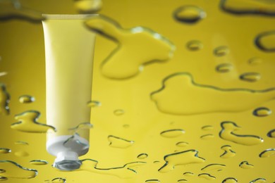 Photo of Moisturizing cream in tube on glass with water drops against yellow background, low angle view. Space for text