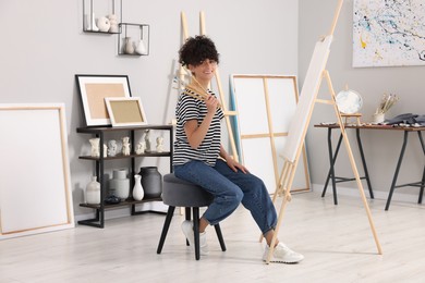 Photo of Young woman holding brushes near easel with canvas in studio