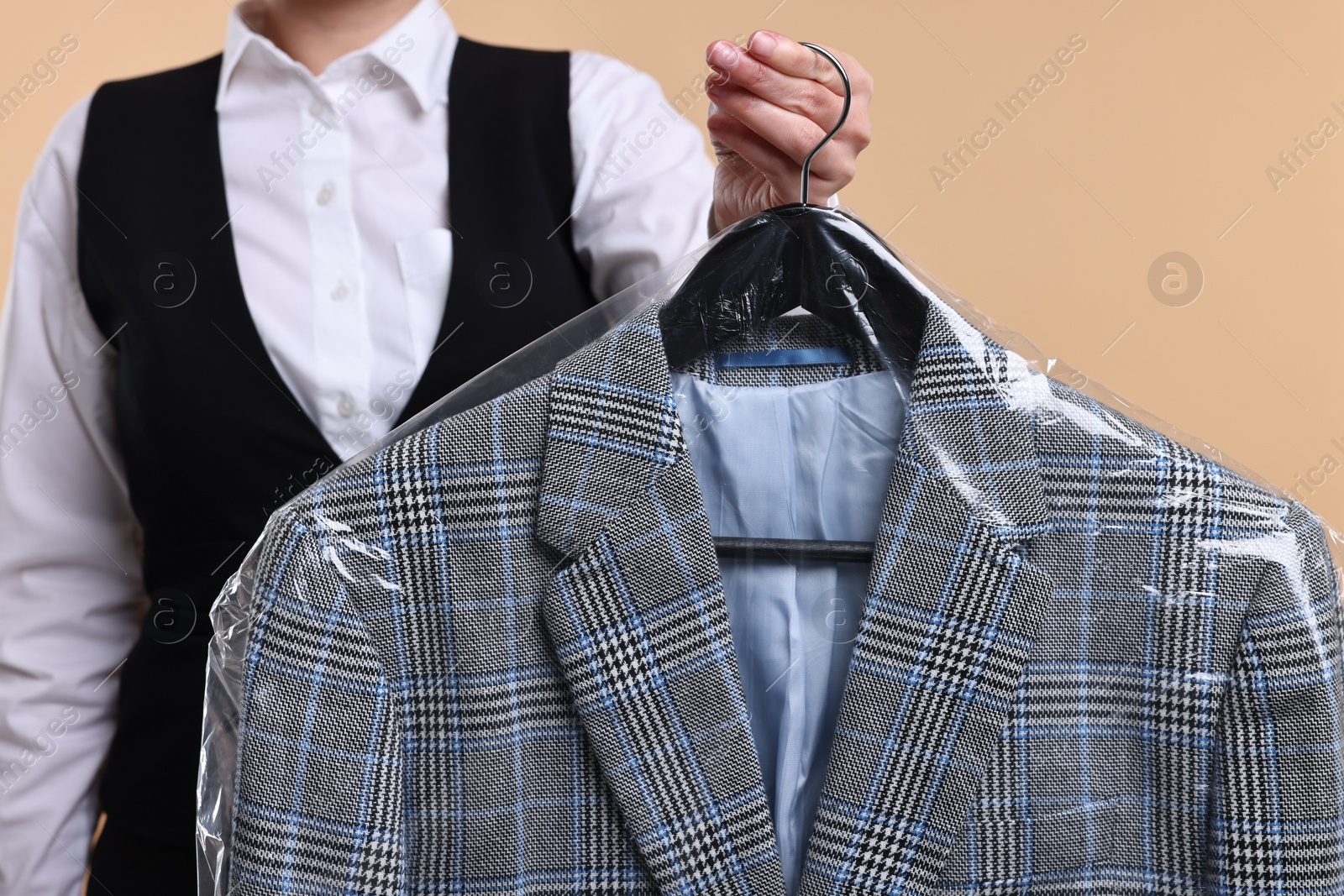 Photo of Dry-cleaning service. Woman holding jacket in plastic bag on beige background, closeup