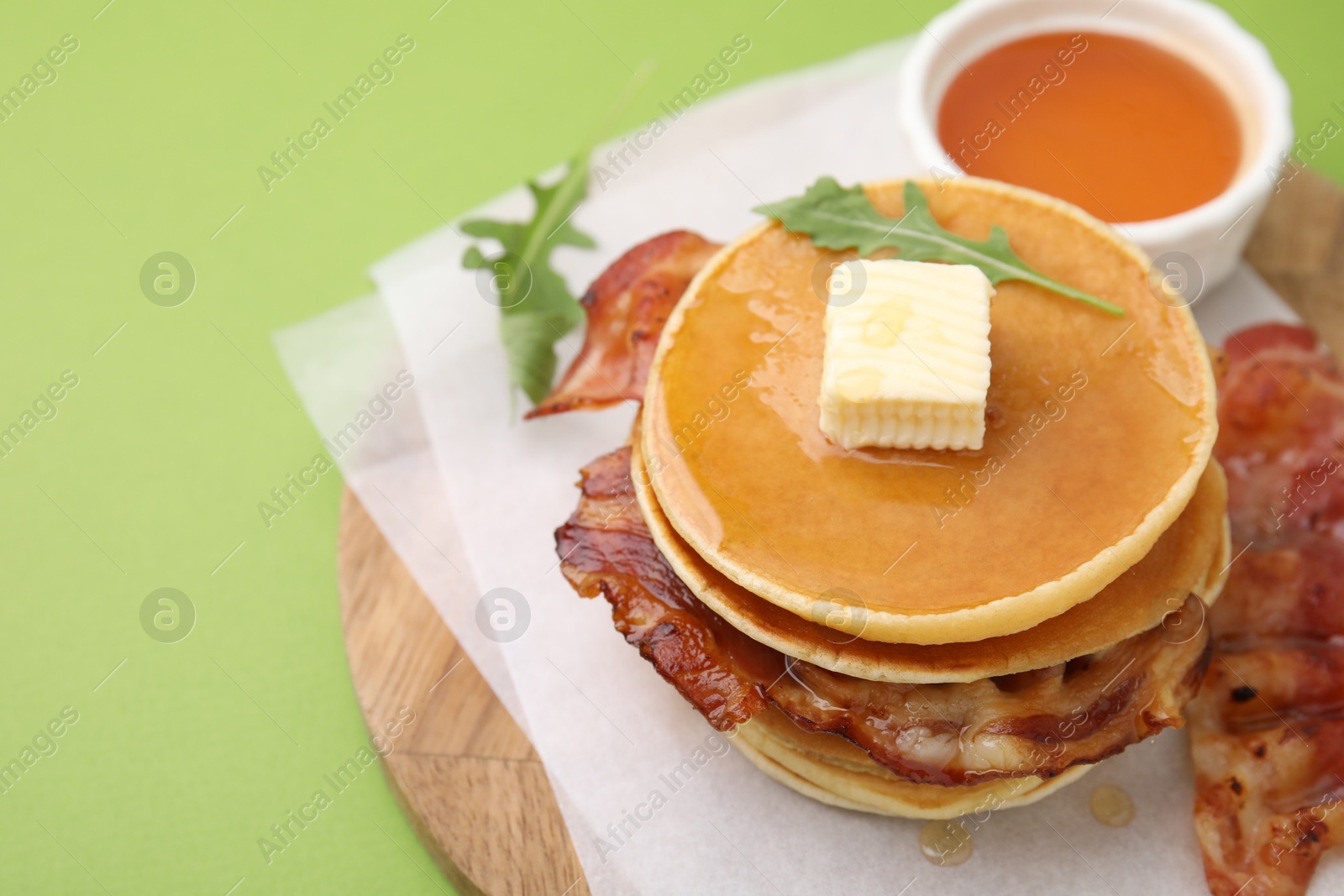 Photo of Delicious pancakes with bacon, butter, arugula and honey on green table, closeup. Space for text