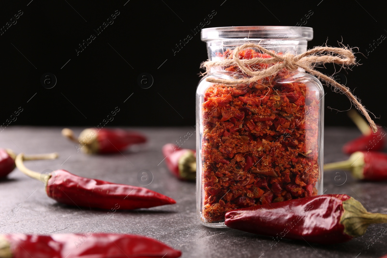 Photo of Chili pepper flakes in jar and pods on textured table against black background, closeup