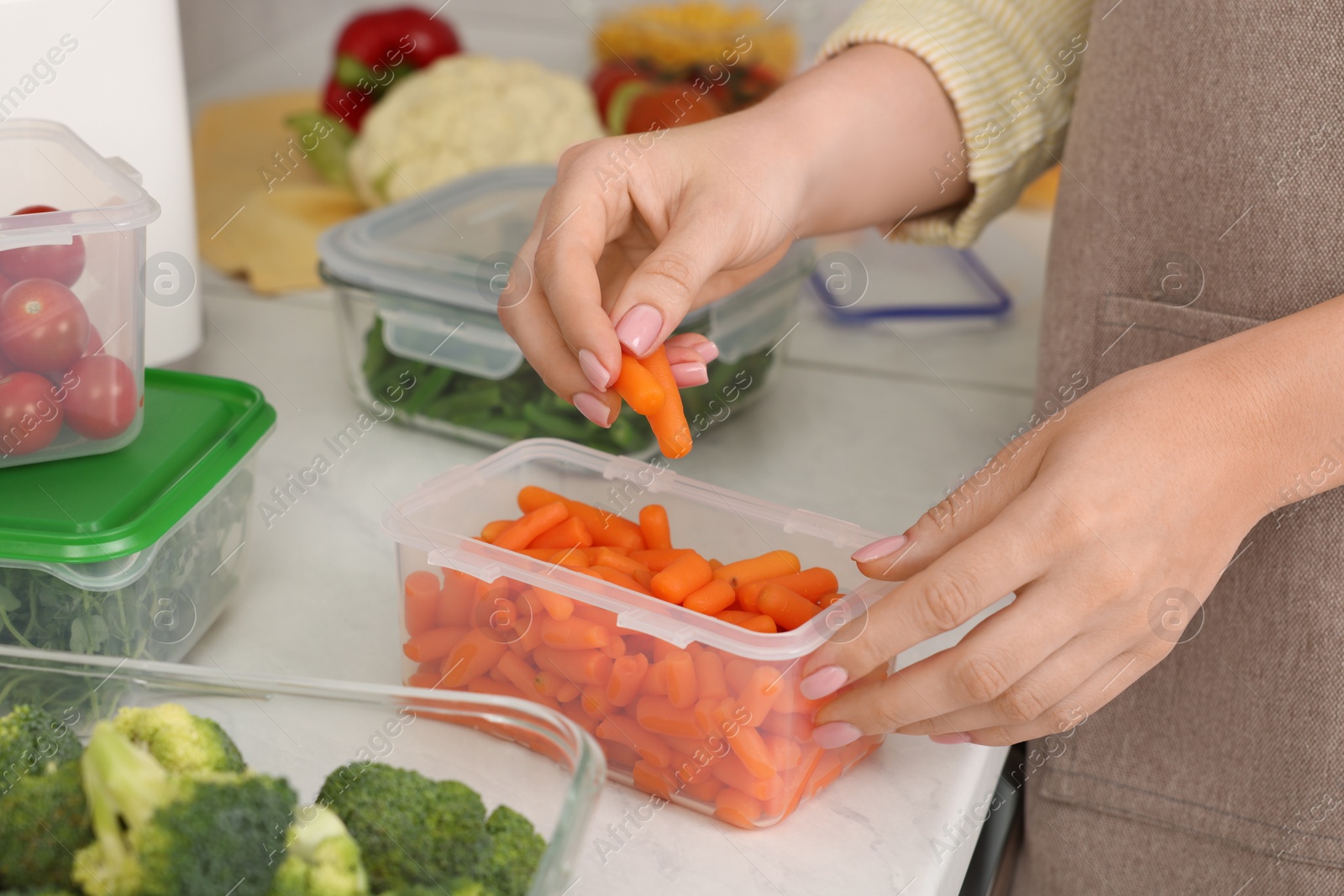 Photo of Woman putting carrots into container with fresh vegetables at white marble table in kitchen, closeup. Food storage