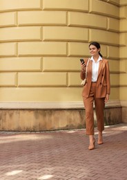 Photo of Young beautiful woman in stylish suit using smartphone near pale yellow building outdoors