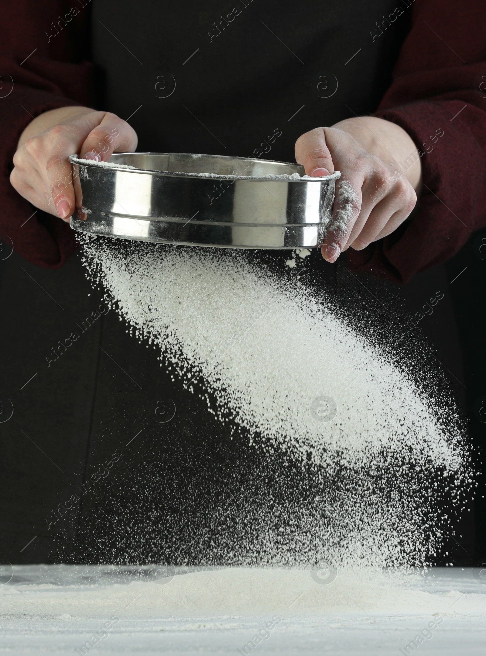 Photo of Woman sieving flour at table against black background, closeup