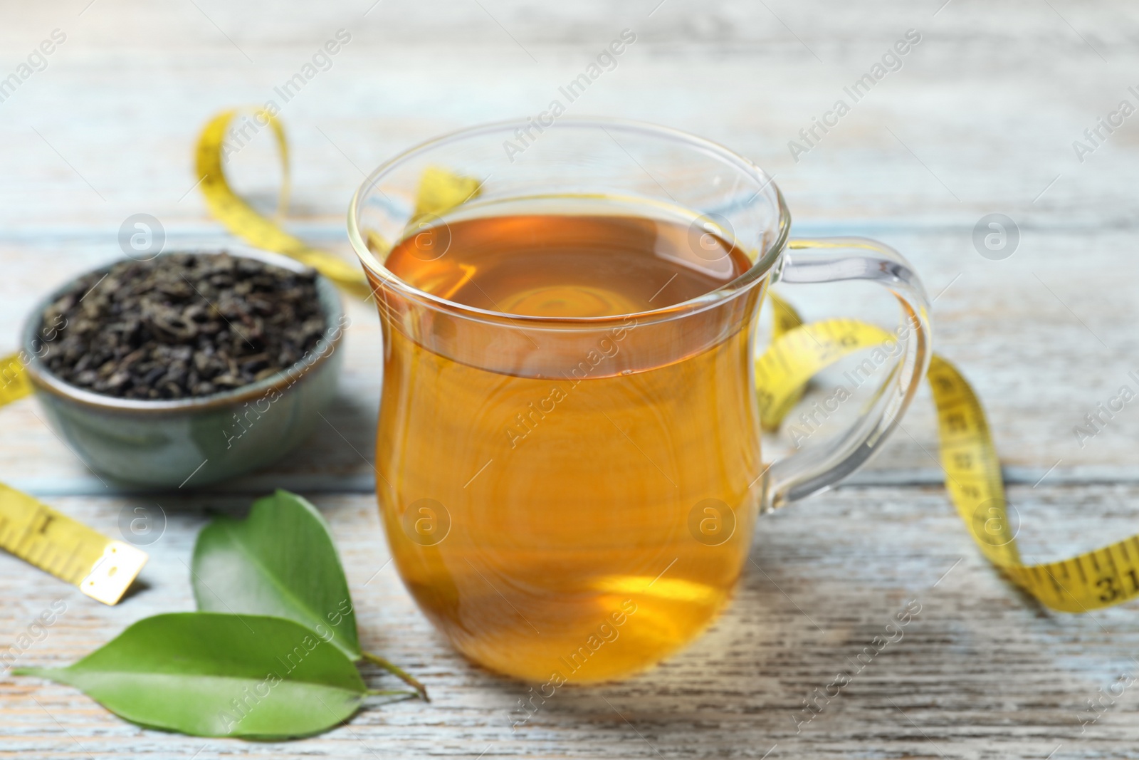 Photo of Cup of herbal diet tea, measuring tape and green leaves on wooden table, closeup. Weight loss concept