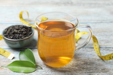 Photo of Cup of herbal diet tea, measuring tape and green leaves on wooden table, closeup. Weight loss concept