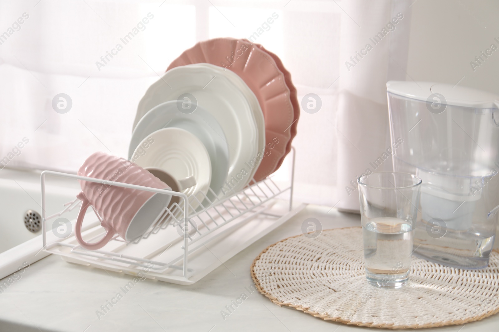 Photo of Drainer with different clean dishware, cup, glass and filter jug on light table indoors