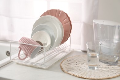 Photo of Drainer with different clean dishware, cup, glass and filter jug on light table indoors