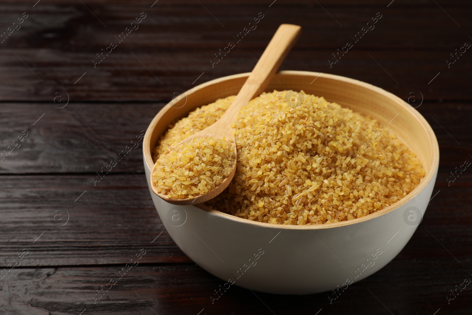 Photo of Bowl and spoon with raw bulgur on wooden table, closeup