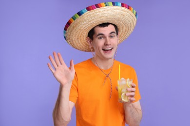 Photo of Young man in Mexican sombrero hat with cocktail waving hello on violet background