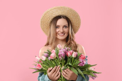 Happy young woman in straw hat holding bouquet of beautiful tulips on pink background