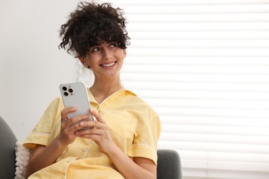 Beautiful young woman in stylish pyjama using smartphone on sofa at home, space for text