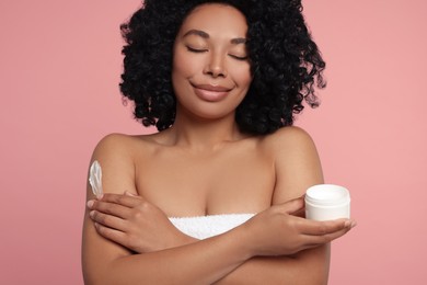 Young woman applying body cream onto shoulder on pink background