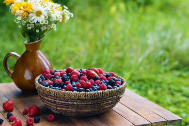 Photo of Wicker bowl with different fresh ripe berries and beautiful flowers on wooden table outdoors, space for text