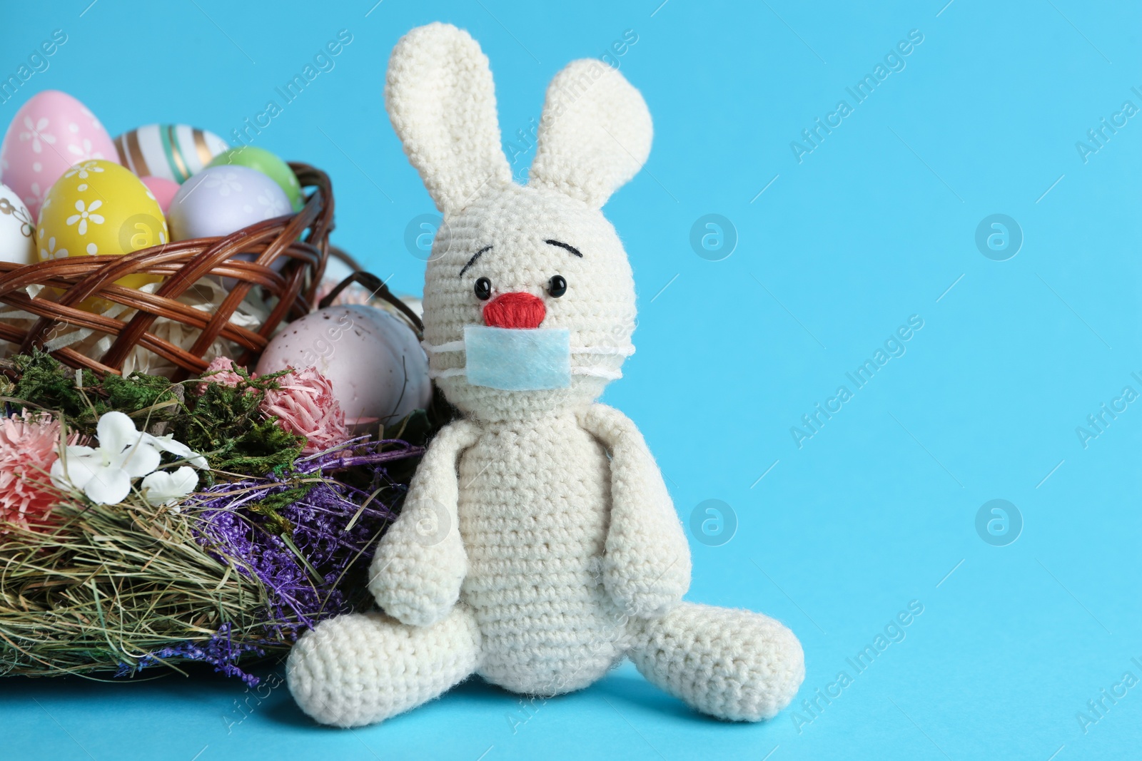 Photo of Toy bunny in protective mask near wicker basket with eggs on light blue background, space for text. Easter holiday during COVID-19 quarantine