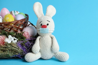 Photo of Toy bunny in protective mask near wicker basket with eggs on light blue background, space for text. Easter holiday during COVID-19 quarantine