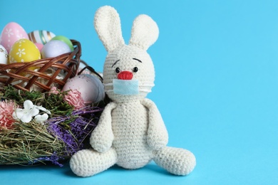 Toy bunny in protective mask near wicker basket with eggs on light blue background, space for text. Easter holiday during COVID-19 quarantine