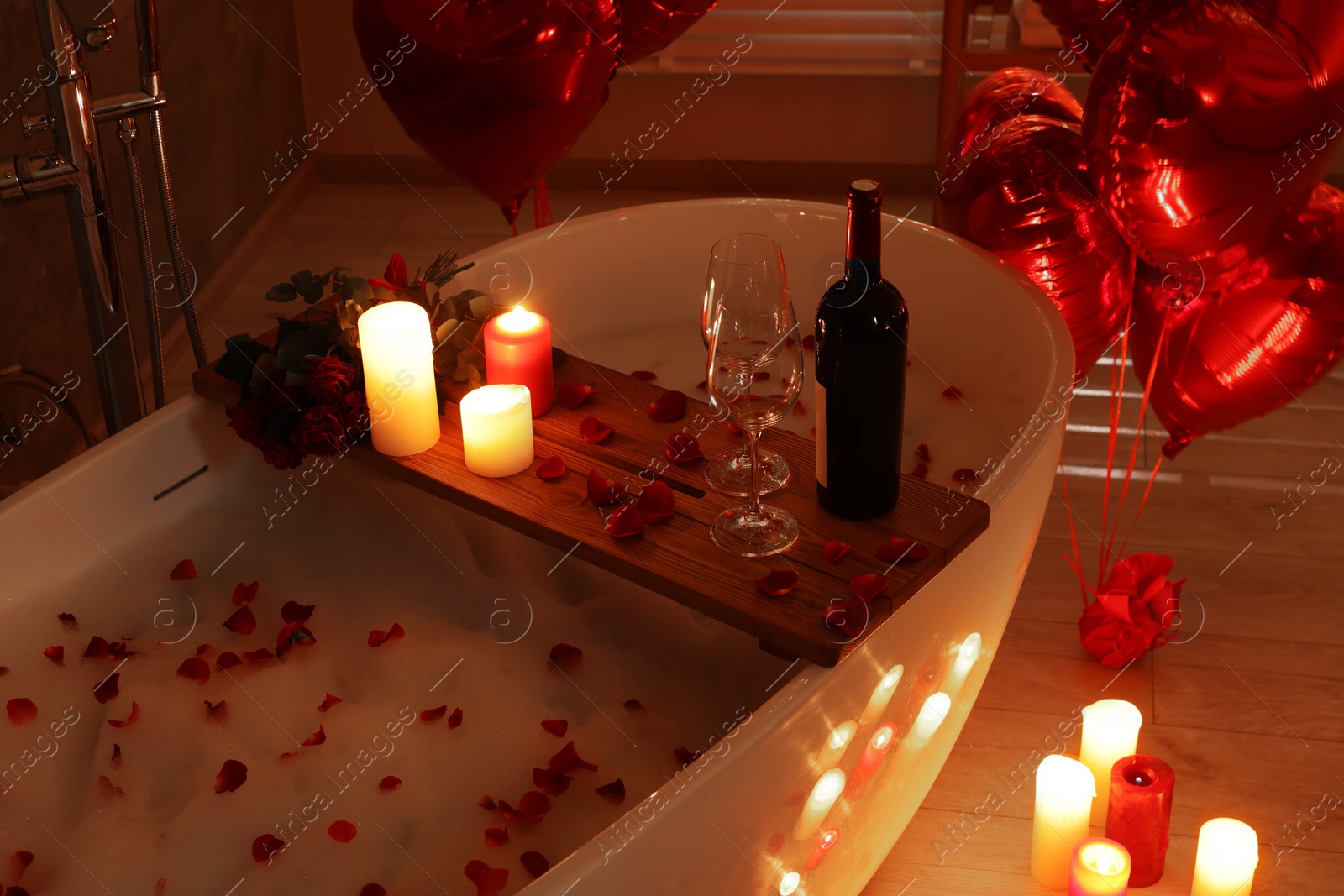 Photo of Wooden tray with wine, burning candles and rose petals on tub in bathroom. Valentine's day celebration