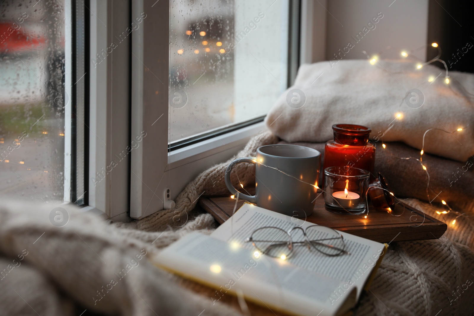 Photo of Cup of hot drink, open book and Christmas lights on knitted blanket near window. Cozy season