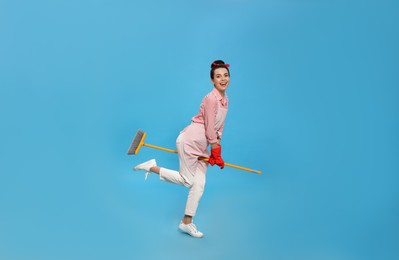 Young housewife with broom having fun on light blue background