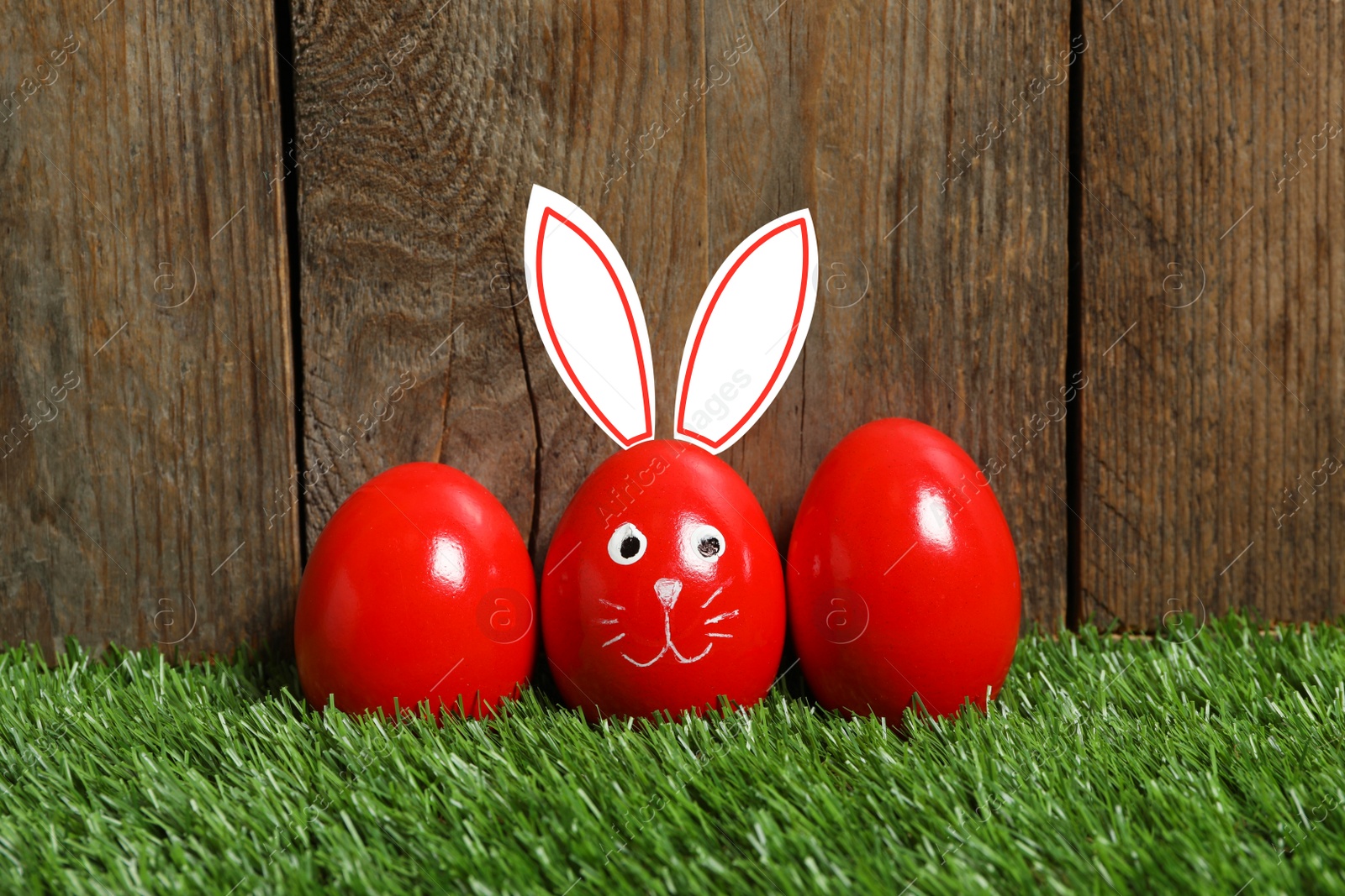 Image of One egg with drawn face and ears as Easter bunny among others on green grass against wooden background