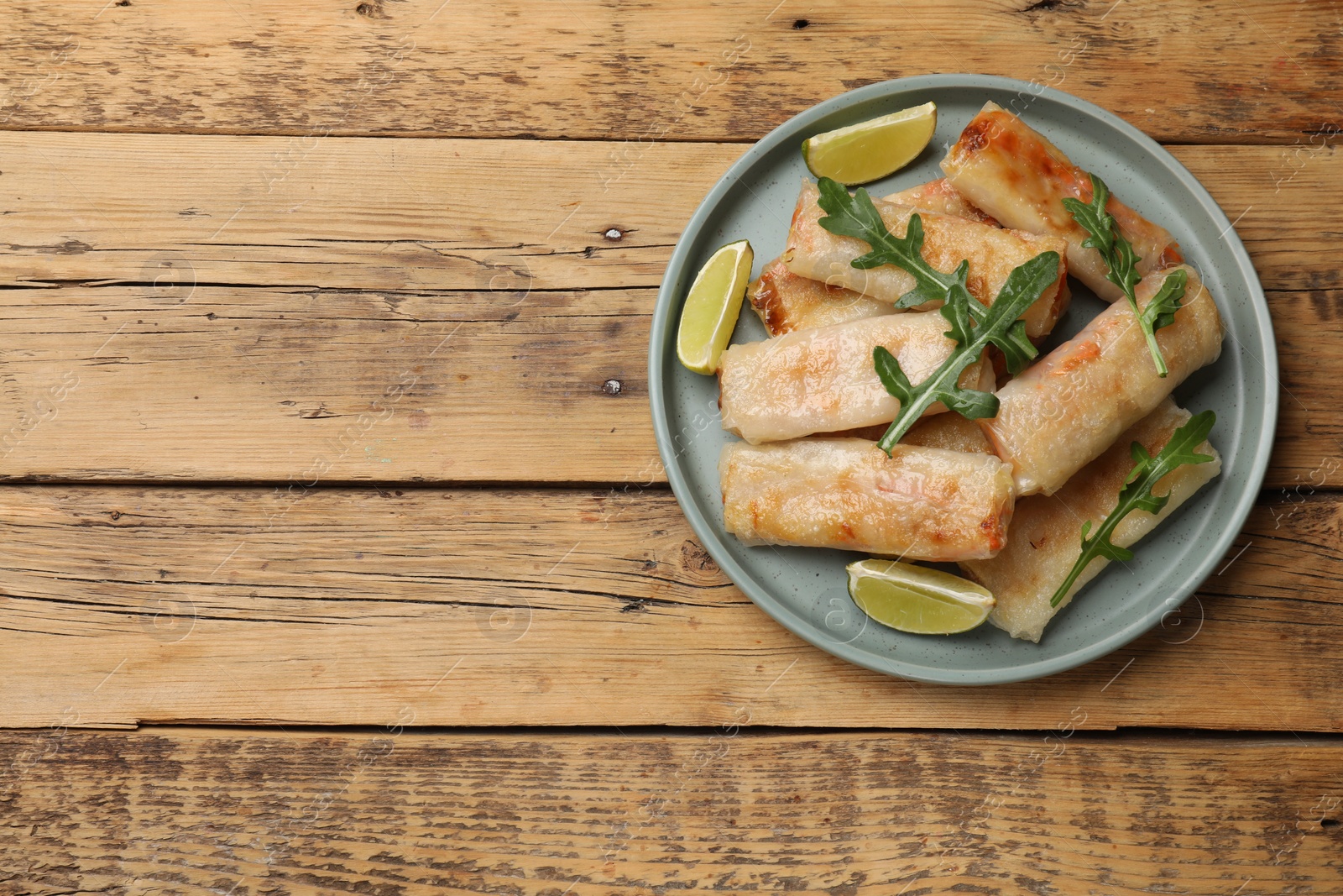 Photo of Plate with tasty fried spring rolls, arugula and lime on wooden table, top view. Space for text