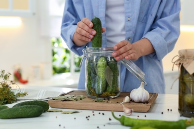 Woman putting cucumber into pickling jar at table in kitchen, closeup