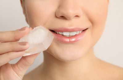 Young woman with ice cube on light background, closeup. Skin care