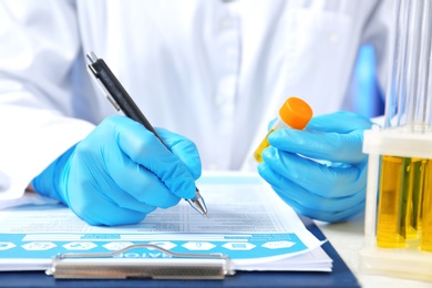 Photo of Laboratory assistant with urine sample for analysis writing medical report at table, closeup