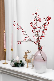 Photo of Hawthorn branches with red berries in vase, candles and deer figures on white table indoors