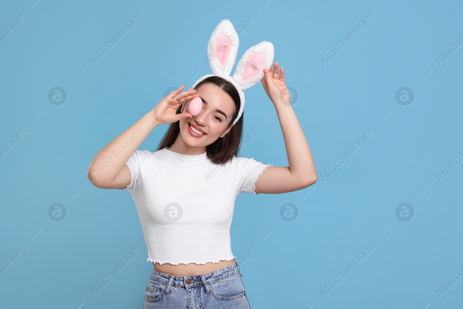 Photo of Happy woman in bunny ears headband holding painted Easter egg on turquoise background